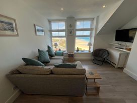 A living room with a sofa and cushions facing two windows overlooking a beach with lamps and a chair next to a mounted TV at Trevose in St Ives