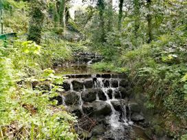 A small stepped waterfall in a forested area with green plants and a metal fence at Llys Madoc The Top Flat in Penmaenmawr