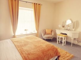 A bedroom with a bed covered in a blanket, a window with curtains, an armchair, and a white dressing table with a mirror at Llys Madoc The Top Flat in Penmaenmawr