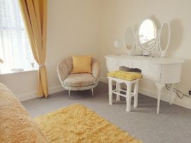 A room with yellow curtains a patterned armchair with a yellow cushion a white dressing table with three mirrors and a yellow cushion on a white stool at Llys Madoc The Top Flat in Penmaenmawr