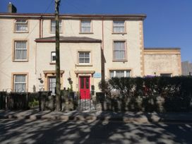 A beige building with multiple windows and a red door with a stone wall and a hedge in front at Llys Madoc The Top Flat in Penmaenmawr