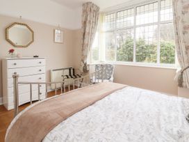 A bedroom with a metal bed frame a white chest of drawers a chair and large windows with floral curtains at Coniston in Rhos Isaf