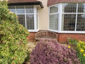 A wooden bench on a patio surrounded by bushes and flowers outside a house with large windows at Coniston in Rhos Isaf