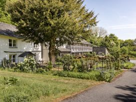 A country house with white walls and a slate roof surrounded by greenery and trees with a fenced garden along a paved road at High Lindeth in Bowness