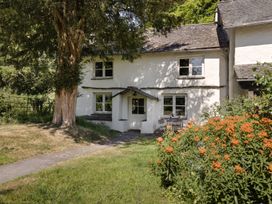 A white cottage with a small porch and windows surrounded by a garden with flowers and a large tree at High Lindeth in Bowness