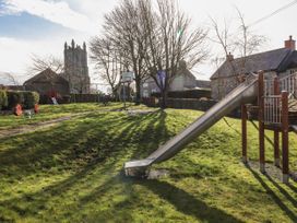 A playground with a slide a basketball hoop swings and a seesaw in a grassy area with stone buildings and a church tower in the background at Northgate Cottage in Ysceifiog near Caerwys