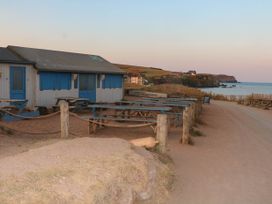 An outdoor seating area with wooden picnic tables next to a blue and white building near a coastal path with a view of cliffs and the sea at 4 Thurlestone Beach House in South Milton near Thurlestone