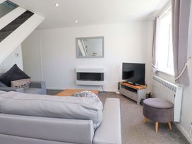 A living room with a grey sofa, armchair, coffee table, wall-mounted electric fireplace, TV on a stand, and a round footstool at Galwad Y Mor in Morfa Nefyn