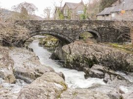 A stone arch bridge over a flowing river with rocky banks and some buildings in the background at Afallon in Penmachno
