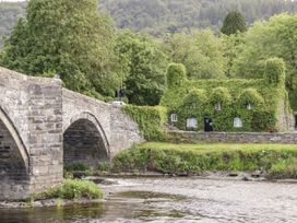A stone bridge over a river with a house covered in greenery in the background at Afallon in Penmachno