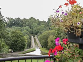 A canal aqueduct surrounded by trees with flowers in the foreground at Belmont Bridge in Henlle