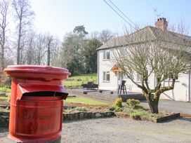 A red postbox with Belmont Bridge written on it in front of a white house with a bare tree and outdoor seating at Belmont Bridge in Henlle