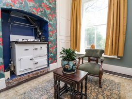 A room with a vintage white stove built into a wall alcove a green upholstered chair a wooden nesting table with a plant a large window with mustard curtains and floral wallpaper at The Woodlands in Berrynarbor