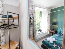 A bedroom with a bed with patterned bedding a bedside table with a lamp a window with curtains and a hanging plant and a wooden shelf with hangers a large wall mirror at The Woodlands in Berrynarbor