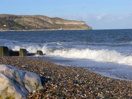 A pebbled beach with waves crashing and a rocky hill in the background with a pier at Pen Y Bont in Llanfair Talhaiarn