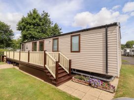 A beige mobile home with a wooden deck and stairs surrounded by grass and trees at 19 The Brambles in Humberston Lincolnshire