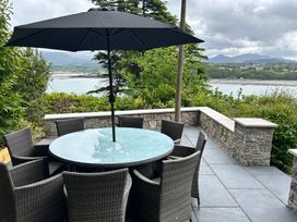 An outdoor patio with a round glass table and wicker chairs under a large umbrella overlooking water and hills at Afallon in Menai Bridge