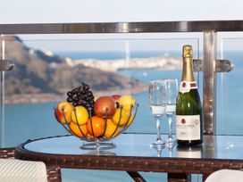 A glass table with a fruit bowl grapes and oranges and a bottle of champagne with two glasses on a balcony overlooking the sea at Harbour View in Carbis Bay
