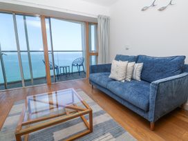 A living room with a blue sofa and glass coffee table facing sliding glass doors opening to a balcony with two chairs and ocean view at Kittiwake in Carbis Bay