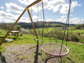 A wooden swing set with two swings and a wooden picnic table on a grassy hill with trees and hills in the background at Fron Goch Farm in Llanfair Talhaiarn
