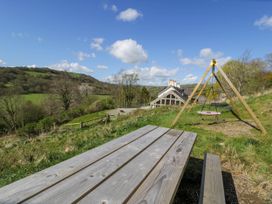 A wooden picnic table and a swing set with a pink circular swing in a grassy area with a house and hills in the background at Fron Goch Farm in Llanfair Talhaiarn