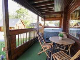 A covered wooden balcony with a round table wooden chairs a hot tub and wooden houses in the background at Sun View Lodge in Llanbedr
