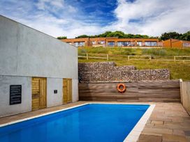 An outdoor swimming pool with wooden doors on a white wall and stone terrace near houses on a grassy hill at Egret in Brixham