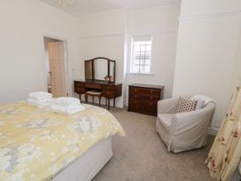 A bedroom with a bed covered in a yellow floral blanket and towels on top a wooden dresser with a mirror a small chest of drawers an armchair and a window at Seawinds 2a in Rhos-On-Sea