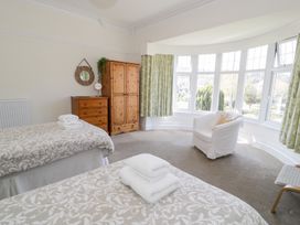 A bedroom with two beds a wooden wardrobe and chest of drawers a white armchair and large bay windows with curtains at Seawinds 2a in Rhos-On-Sea