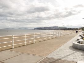 A paved promenade with white railings along a sandy beach with hills and buildings in the distance at Seawinds in Rhos-On-Sea