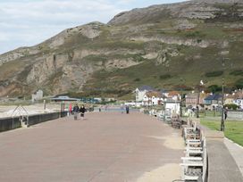 A seaside promenade with benches along the right side and several people walking with hills in the background at Seawinds in Rhos-On-Sea