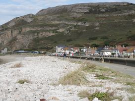 A rocky shoreline with a paved walkway and people walking near houses and grassy hills at Seawinds in Rhos-On-Sea