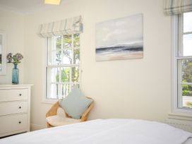 A bedroom with a wicker chair with cushions next to two windows a white chest of drawers with a vase of flowers and a painting on the wall at Lowenna Manor 3 in Rock