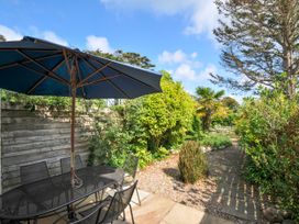 An outdoor patio area with a black metal table and chairs under a large blue umbrella next to a wooden fence and a garden path surrounded by various plants and trees at Lowenna Manor 3 Rock
