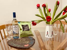 A glass table with a bottle of white wine, two wine glasses, a packet of vegetable crisps, a note on a stone, and a white jug with red flowers at 7 Watkin Street in Conwy