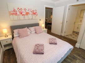 A bedroom with a double bed and pink bedspread with two pink pillows and folded towels two bedside tables with lamps a flamingo painting on the wall and open doors leading to a dining room and a bathroom at Rookley Lake Cottage in Rookley near Godshill