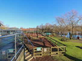 An outdoor play area with wooden climbing structures and a fenced section near a lake with trees and picnic tables at Rookley Lake Cottage in Rookley near Godshill