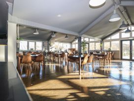 A dining area with multiple tables and brown chairs in a room with large windows and hanging lights at Rookley Lake Cottage in Rookley near Godshill