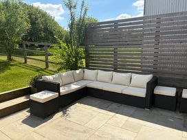 An outdoor patio area with a black wicker sectional sofa with white cushions and a wooden slatted fence with grass and trees behind at The Gully in Sherborne