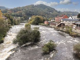A river with rushing water flowing between trees and buildings in a hilly area at Cae Iocyn in Chirk
