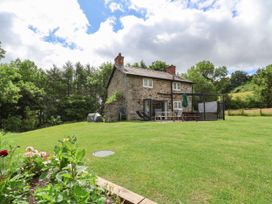 A stone cottage with a chimney and outdoor furniture on a grassy lawn surrounded by trees at Cae Iocyn in Chirk