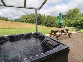 An outdoor patio with a hot tub wooden picnic table with a green umbrella and glass table with two chairs overlooking a grassy field and trees at Cae Iocyn in Chirk