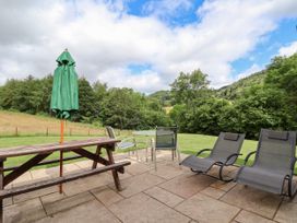 An outdoor patio with a wooden picnic table with a green umbrella two lounge chairs and a round glass table with chairs surrounded by grass and trees at Cae Iocyn in Chirk