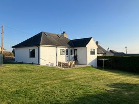 A white house with a black roof and outdoor wooden table with chairs on a patio surrounded by a grassy lawn at Angorfa in Aberdaron