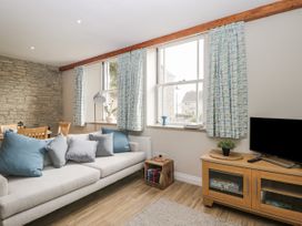 A living room with a sofa and cushions next to three windows with patterned curtains and a wooden TV stand with a plant at The Old Haberdashery in Swanage