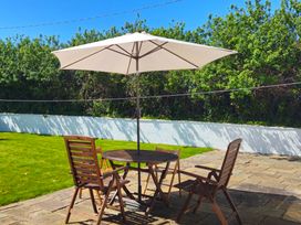 A patio with a wooden table and four chairs under a large white umbrella surrounded by grass and bushes at 3 Tai Newydd in Llanfaelog near Rhosneigr