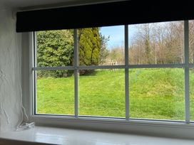 A window with a black blind looking out to a garden with green grass trees and bushes at Min Y Mor in Llanystumdwy near Criccieth