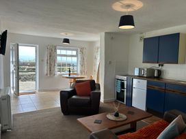 A small living room with a brown chair a sofa and coffee table next to a kitchen with blue cabinets and a dining table near windows and an open door at Min Y Mor Llanystumdwy near Criccieth