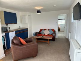 A living room with a brown armchair and a brown sofa with cushions a wooden coffee table a blue kitchenette and a doorway leading to a bedroom at Min Y Mor in Llanystumdwy near Criccieth