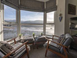 A sitting area with two armchairs, two ottomans, small plants on the window sill, and a view of the water and hills at Harbour View - Flat 2 in Barmouth